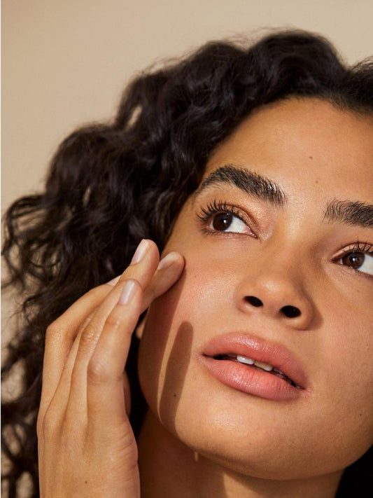 Close-up of a woman with curly hair touching her face showing sensitive skin, against a neutral background